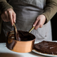 Two hands stirring a copper pot of melted chocolate being used to top a tart